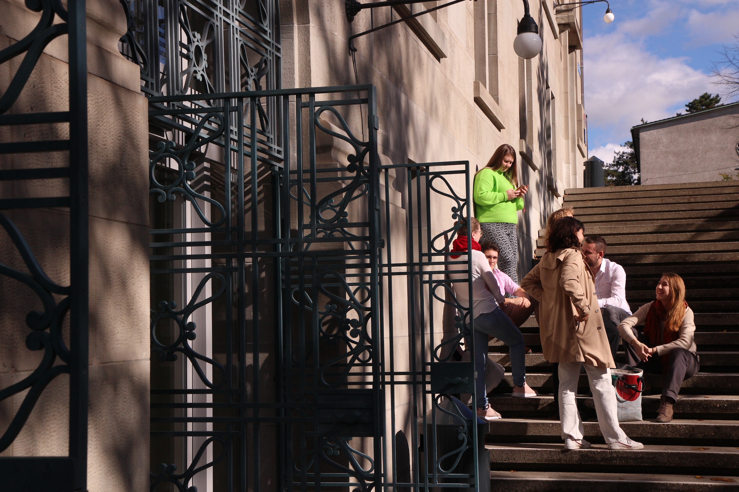 Das Bild zeigt ein einen Eingang des Hauptgebäudes der Universität, eine Gruppe von Studierenden stehen und sitzen auf der Freitreppe und diskutieren.