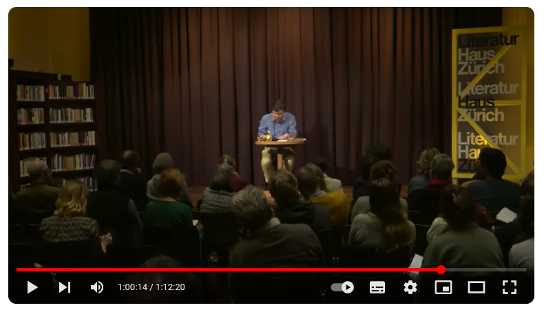 Auf dem Foto sitzt - ganz hinten - Peter Stamm in seinem typischen blauen Hemd an einen kleinen Tisch und liest dem Publikum vor. Im Vordergrund sind sitzende Menschen von hinten zu sehen. Es ist dunkel im Saal.