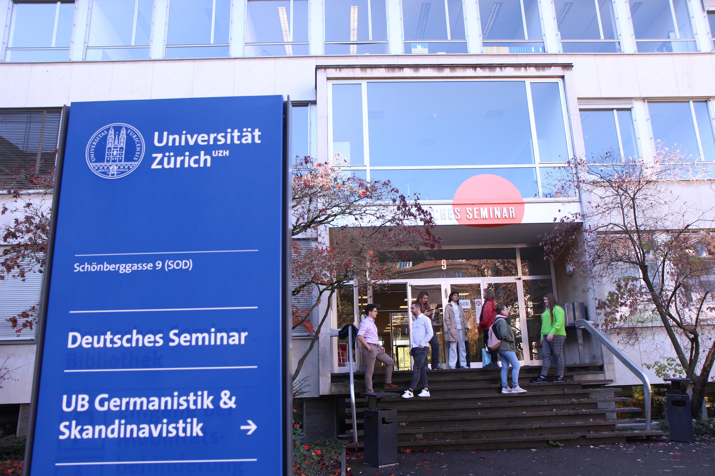 Das Bild zeigt das Deutsche Seminar mit Blick auf den Haupteingang, die grosse Glasfront, die Freitreppe, auf der Studierende stehen und diskutieren. Im Vordergrund steht die Hinweistafel im typischen Blau der Universität.