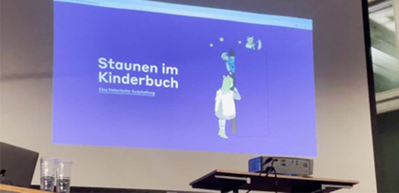 Das Bild zeigt eine Projektionsleinwand hinter einem Beamer. Aus der Leinwand ist die Startseite der Online-Ausstellung "Staunen im Kinderbuch" in sattem Blau zu sehen.