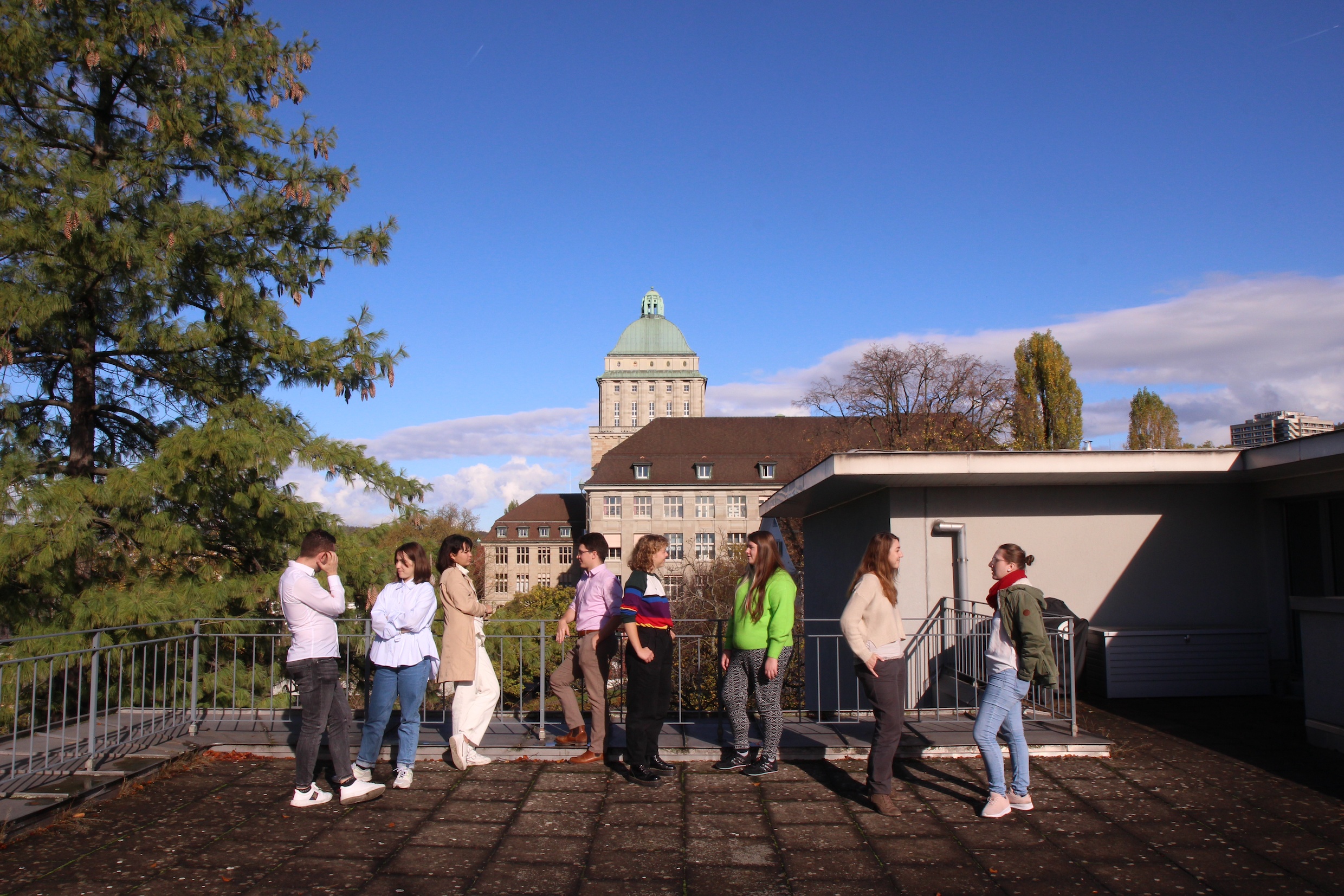 Das Bild zeigt eine Gruppe von Studierenden auf der Dachterrasse des Deutschen Seminars im Gespräch. Im Hintergrund ist der markante Turm des Hauptgebäudes der Universität zu sehen, darüber ein strahlend blauer Himmel.