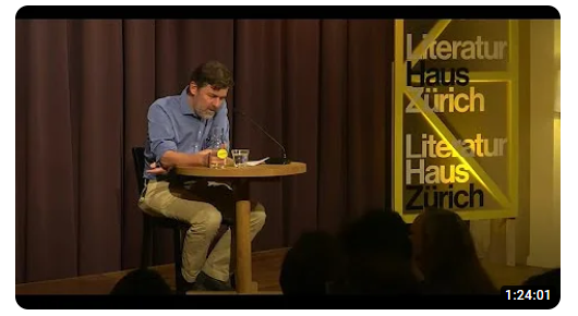 Das Foto zeigt Peter Stamm in einem blauen Hemd an einem kleinen runden Tisch auf der Bühne im Literaturhaus. Es liest gebückt über seinen Text vor.