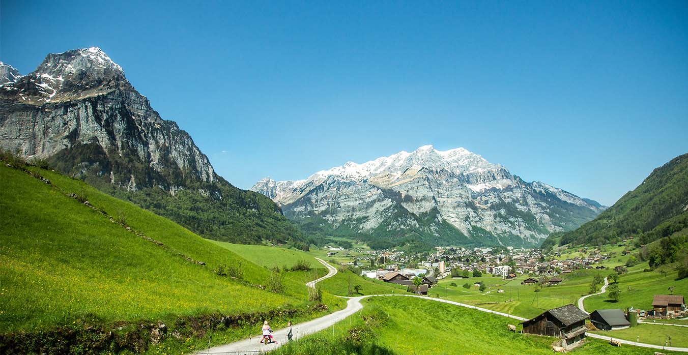 Das Bild zeigt eine Sommerlandschaft, grüne Matten mit Blick auf Glarus, im Hintergrund schneebedeckte Berge.