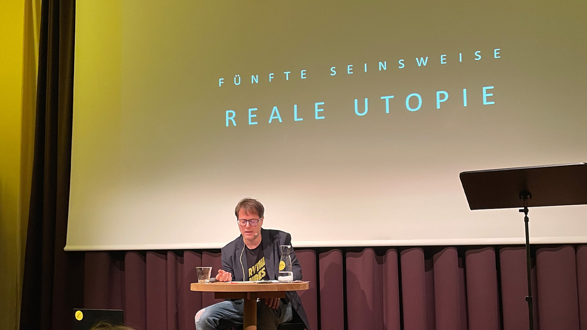 Das Bild zeigt Milo Rau lesend auf der Bühne im Literaturhaus Zürich. Im Hintergrund ist eingeblendet: "Fünfte Seinsweise. Reale Utopie"