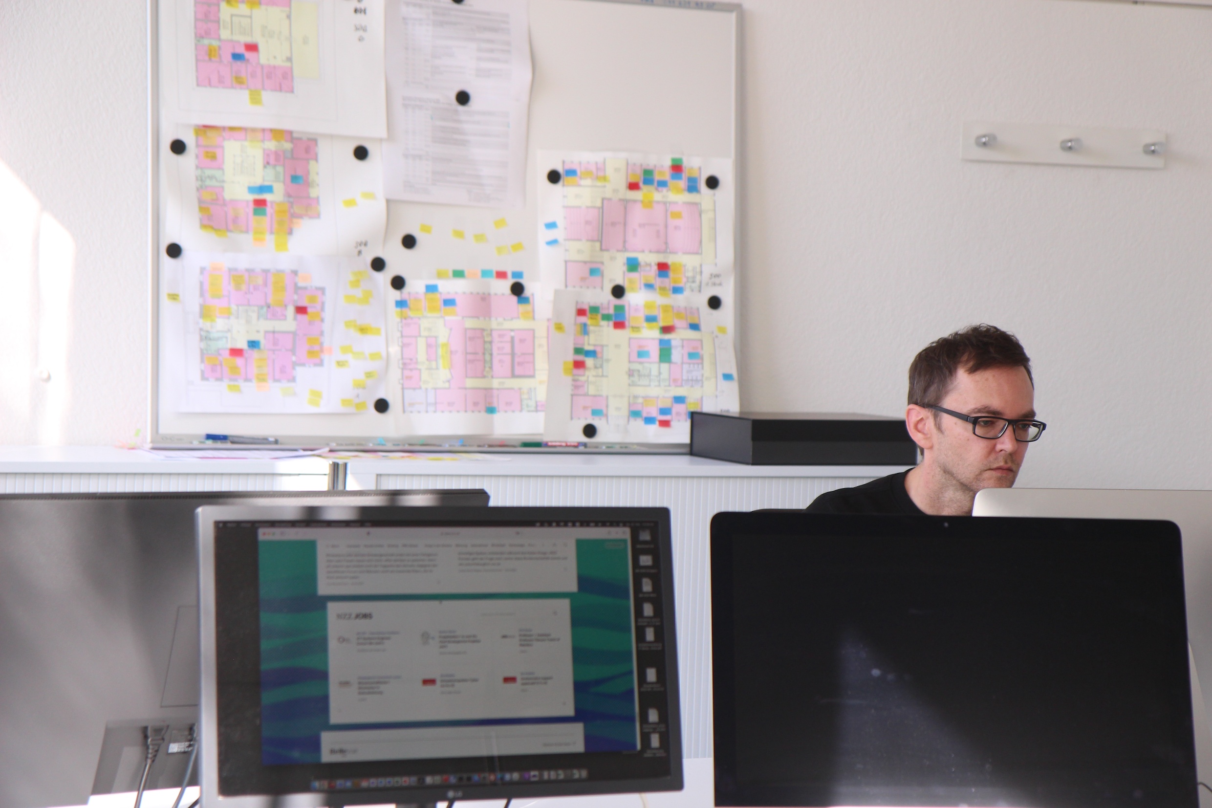 Das Bild zeigt einen Mitarbeiter der IT am Deutschen Seminar in seinem Büro, er sitzt hinter einem Bildschirm. Die untere Hälfte des Bilds ist voll von Bildschirmen, im Hintergrund hängt ein Gebäudeplan.