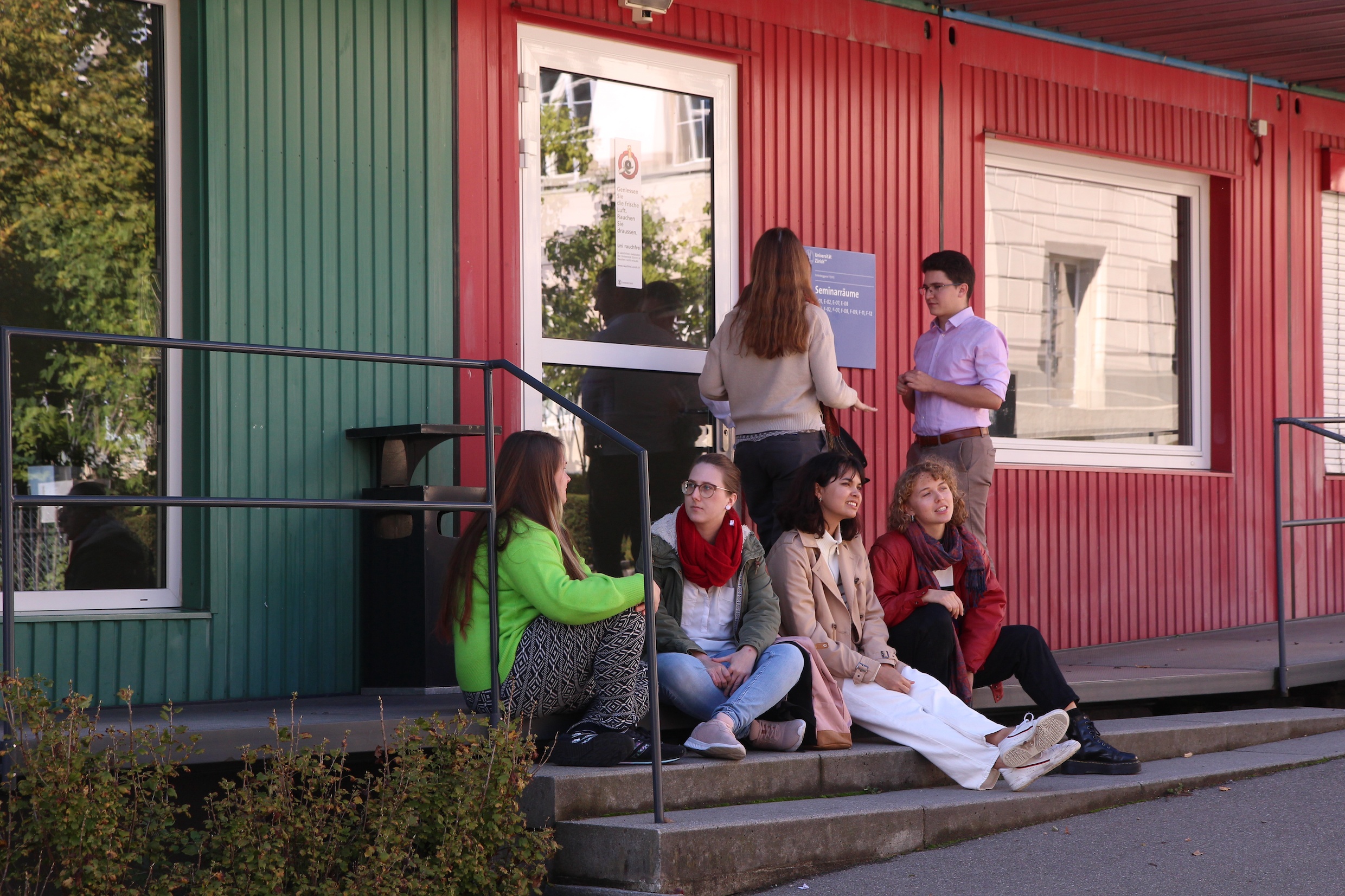 Das Bild zeigt eine Gruppe von Studierenden, die vor einem Gebäude der Universität in rot-grün sitzen und stehen, sie unterhalten sich.