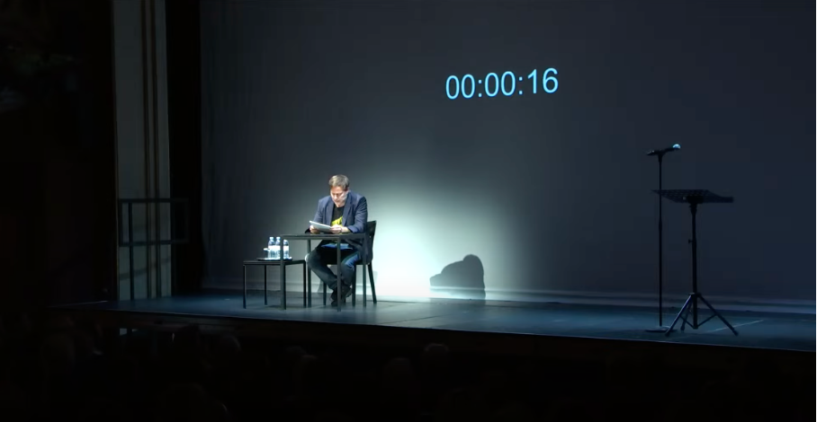 Das Bild zeigt Milo Rau auf der Bühne des Zürcher Schauspielhauses zu Beginn seiner Poetikvorlesung. Die Bühne ist dunkel, ein Scheinwerfer ist auf Milo Rau gerichtet, der an einem kleinen Tisch sitzt und liest.